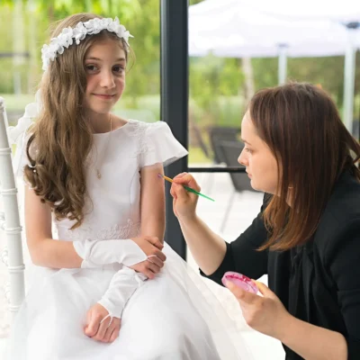 Młoda dziewczyna w białej sukience i kwiecistej opasce siedzi, podczas gdy kobieta nakłada farbę na jej ramię. Dziewczyna uśmiecha się delikatnie. Są w pomieszczeniu z dużymi oknami i zielenią widoczną na zewnątrz. - Kubiccy Fotografia - Fotograf Łódź