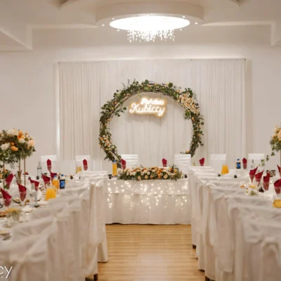 Elegancka sala weselna z długimi stołami nakrytymi białą pościelą, czerwonymi serwetkami i kwiatowymi dekoracjami. Z przodu, okrągły kwiatowy łuk ze światłami i napisem Kubiccy zdobi główny stół. - Kubiccy Fotografia - Fotograf Łódź