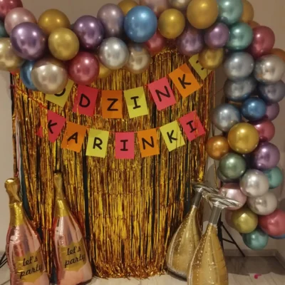 Świąteczne tło imprezy z błyszczącymi złotymi zasłonami z frędzlami, wielokolorowymi balonami ułożonymi w łuk i banerami z napisem DZINKI KARINKI. Cztery nadmuchiwane butelki z napisem lets party na dole. - Kubiccy Fotografia - Fotograf Łódź